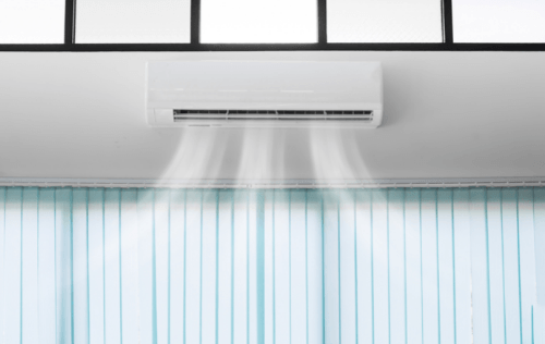 Air conditioning unit mounted on the wall with cool air blowing out, adjacent to light blue vertical blinds, illustrating indoor climate control solutions for minimizing heat.