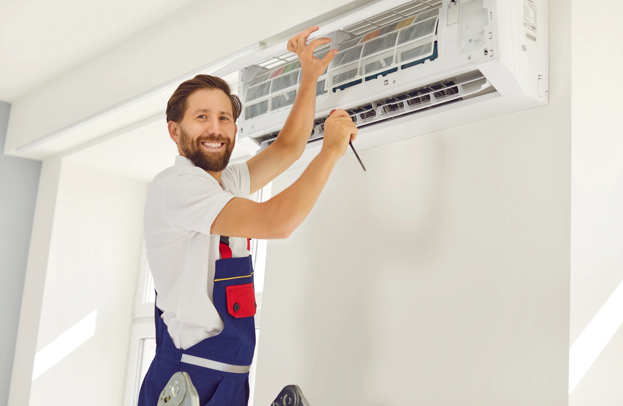 A smiling technician in overalls fixes an air conditioner on a white wall, holding a screwdriver. 