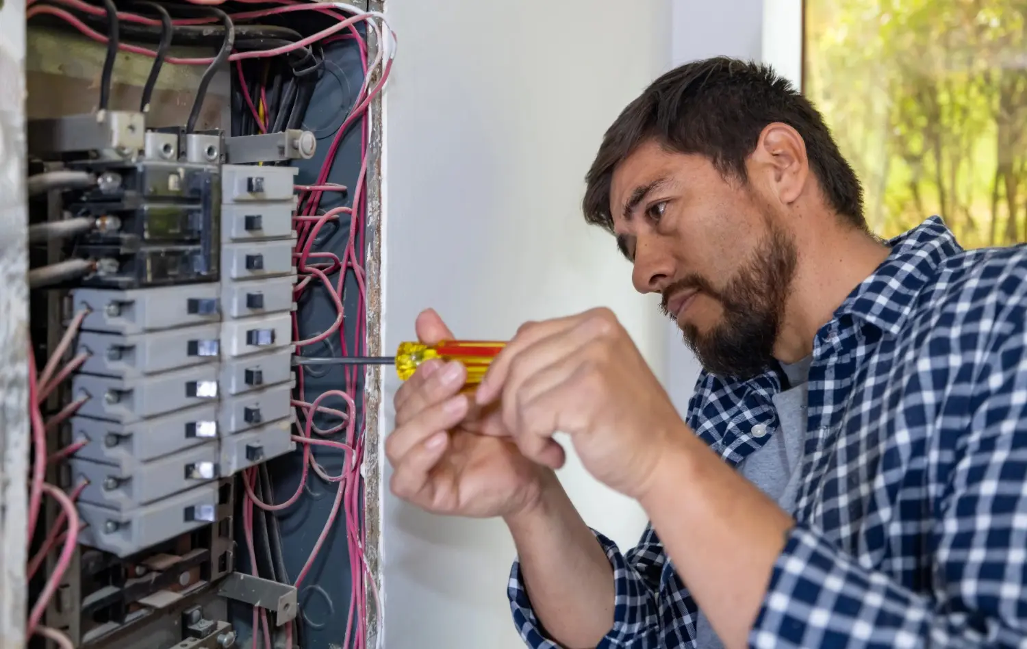 An electrician in a plaid shirt carefully uses a yellow screwdriver to adjust a circuit breaker