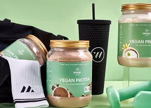 Two jars of vegan protein powder with green labels, a black textured tumbler with a straw, and light green dumbbells on a light green surface.