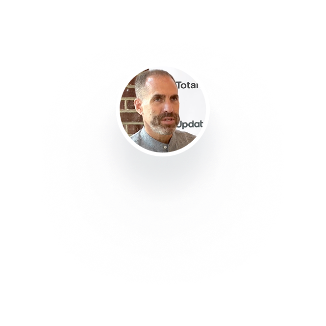 Portrait of a man with a trimmed beard and short hair wearing a buttoned gray shirt, in front of a white background with partial text and a brick wall.