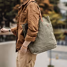 Person wearing a brown jacket and beige pants carrying a large gray tote bag outdoors.