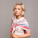 Blonde woman with short hair wearing a white shirt and colorful striped scarf tied around her shoulders against a plain background.