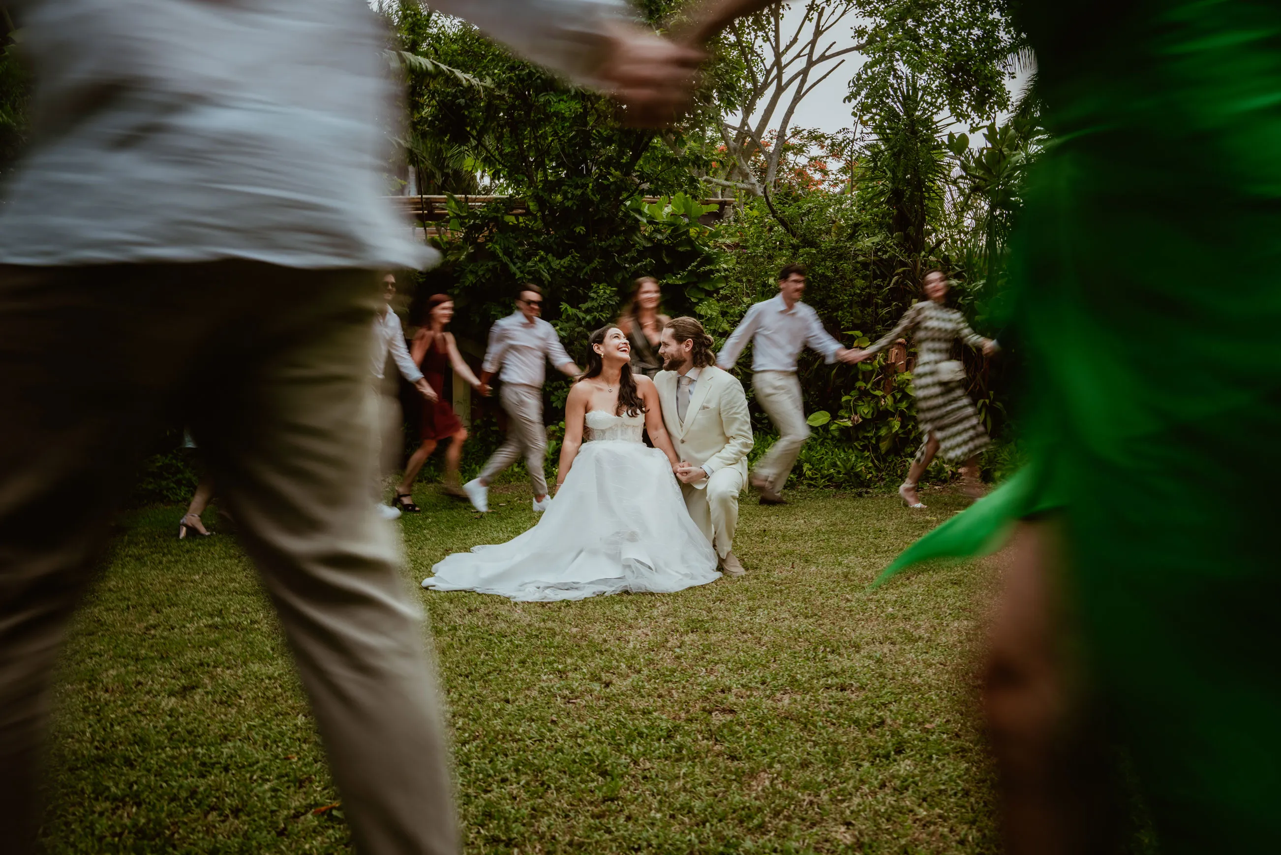 Novios sentados en el jardín rodeados de invitados tomados de la mano