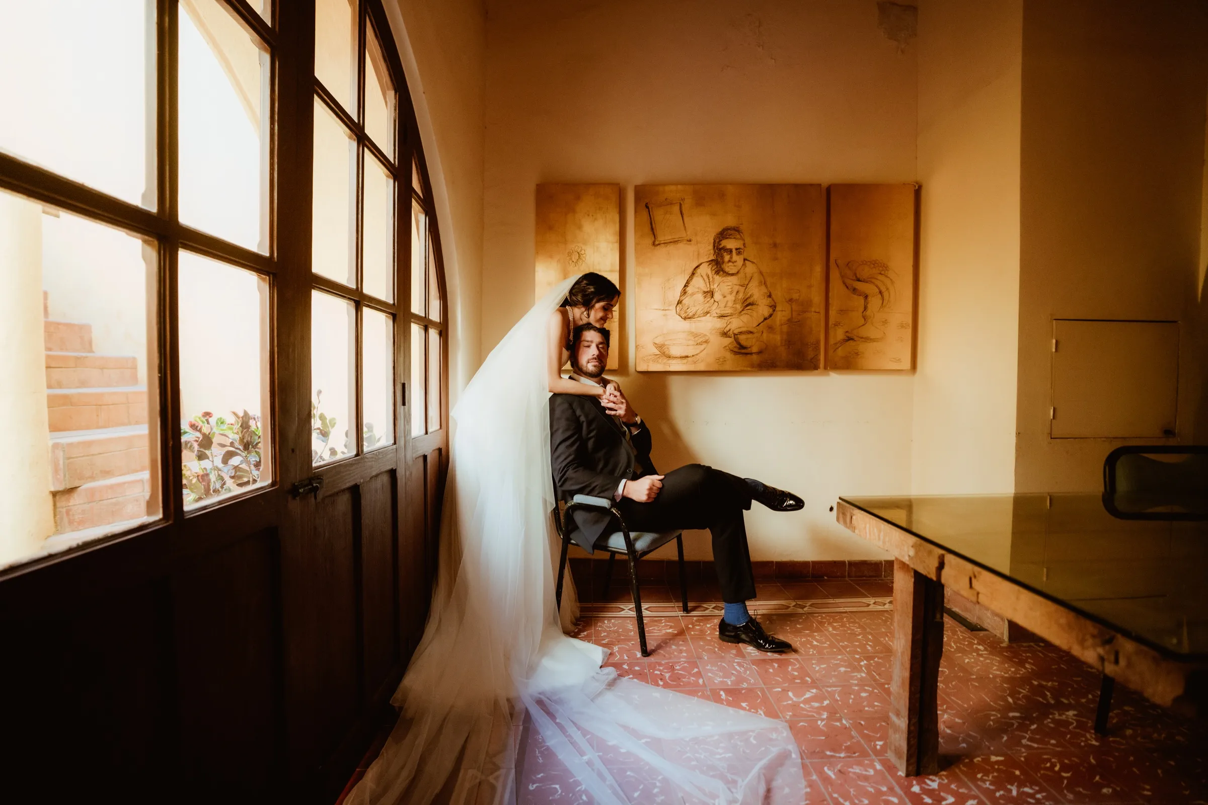 Retrato de boda de la novia detrás del novio sentado junto a una ventana
