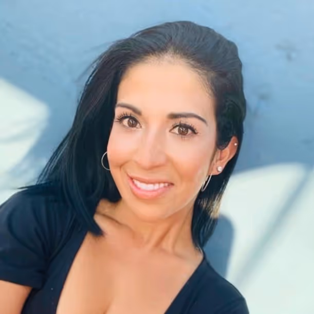 Smiling woman with straight black hair, wearing hoop earrings and a black top against a blurred light blue background.