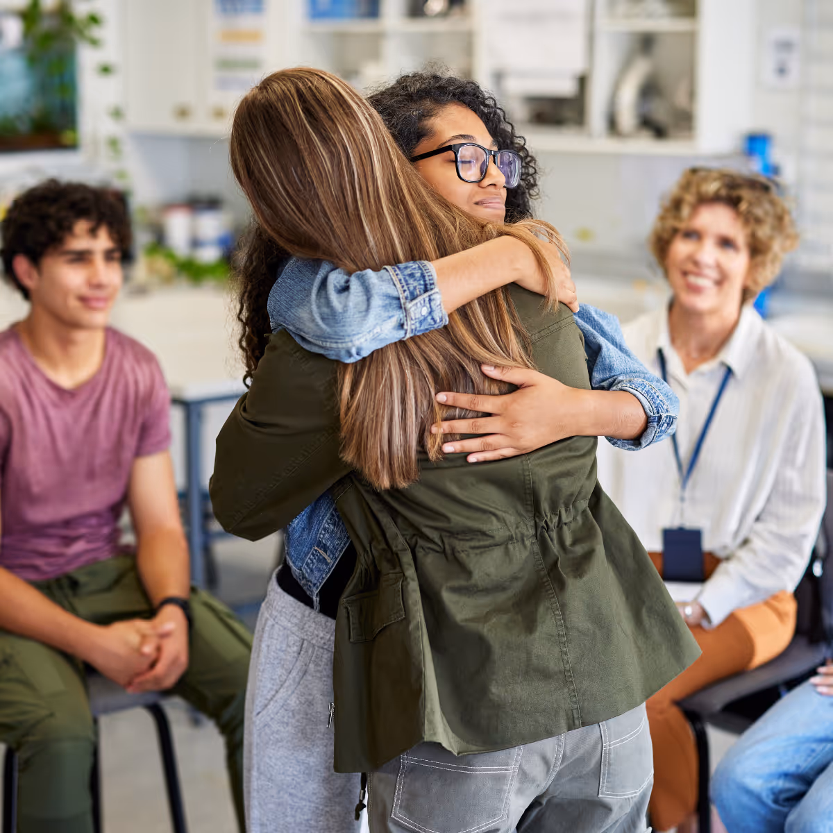 Two young women hugging warmly while others sit watching and smiling in a casual indoor setting.