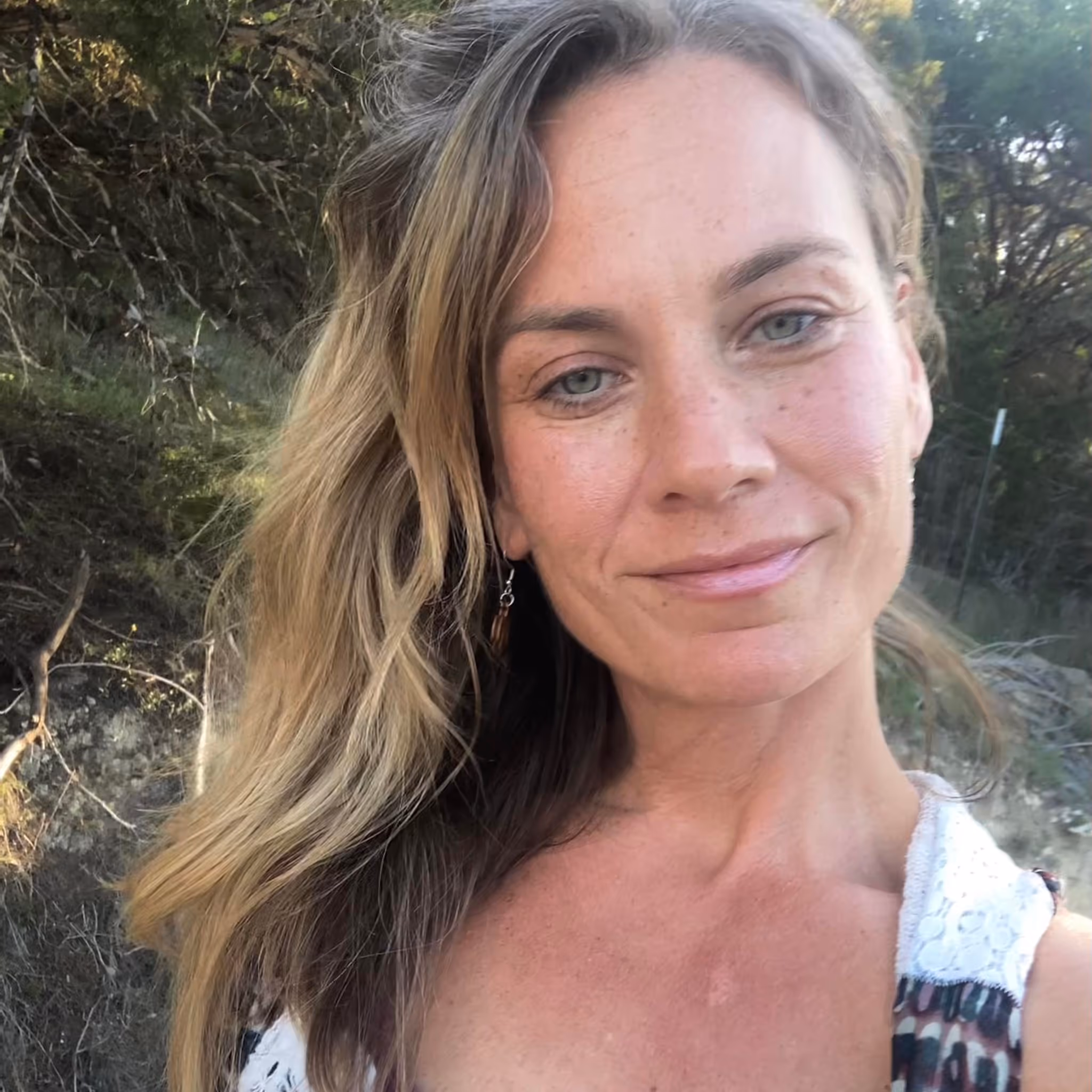 Close-up of a smiling woman with wavy blonde hair standing outdoors with trees in the background.