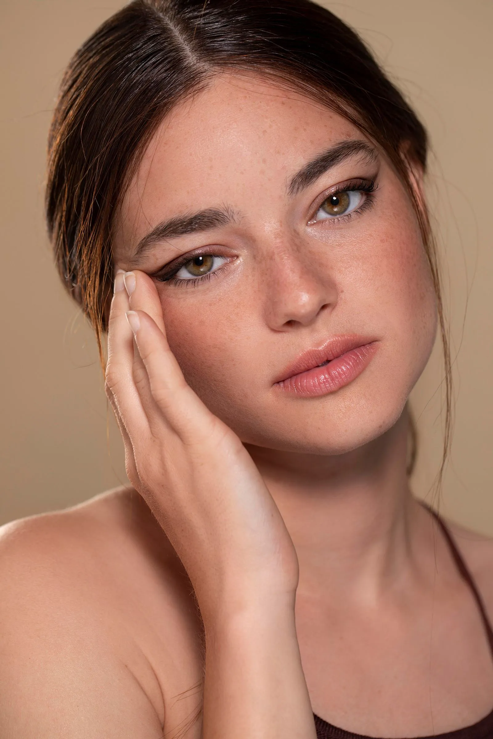 Close-up portrait of a young woman with green eyes, natural makeup, and dark hair, gently touching her face, against a neutral beige background