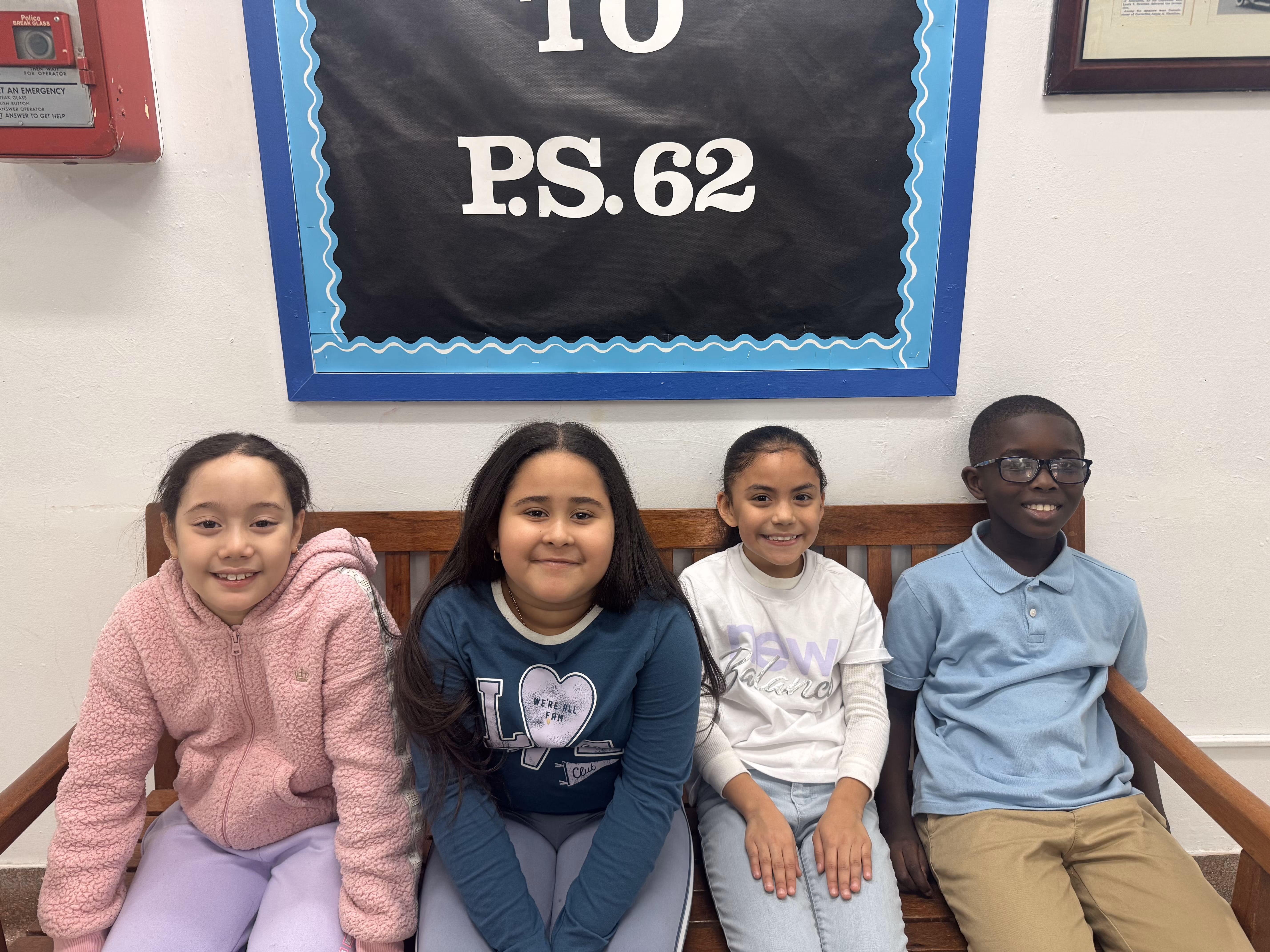 Four smiling PS 62 students seated on a bench under school sign