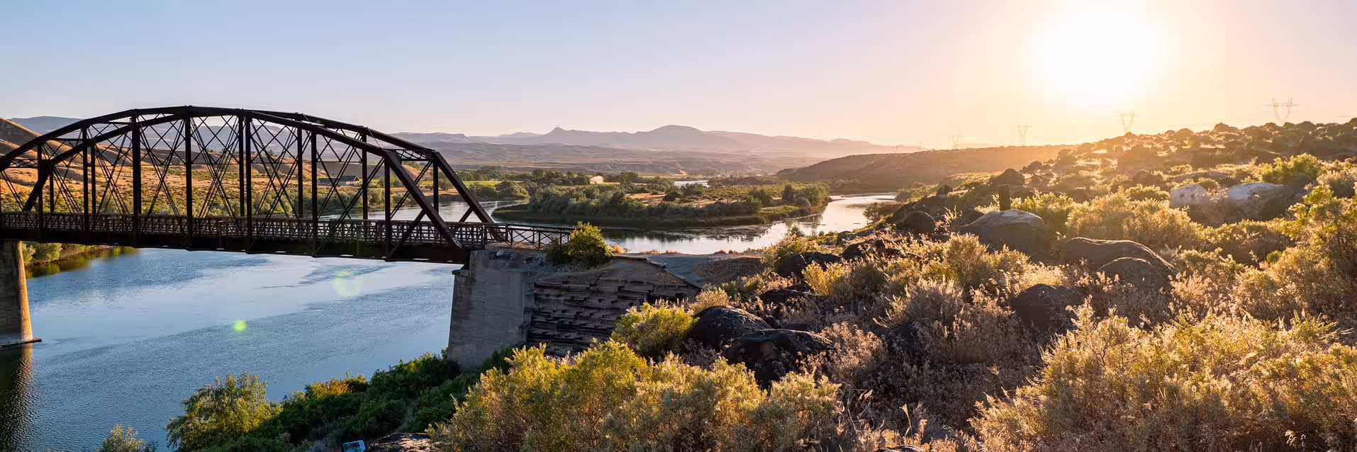 Sunset panorama at Celebration Park, Idaho — bridge with foreground shrubs and silhouetted power lines under an orange sky.