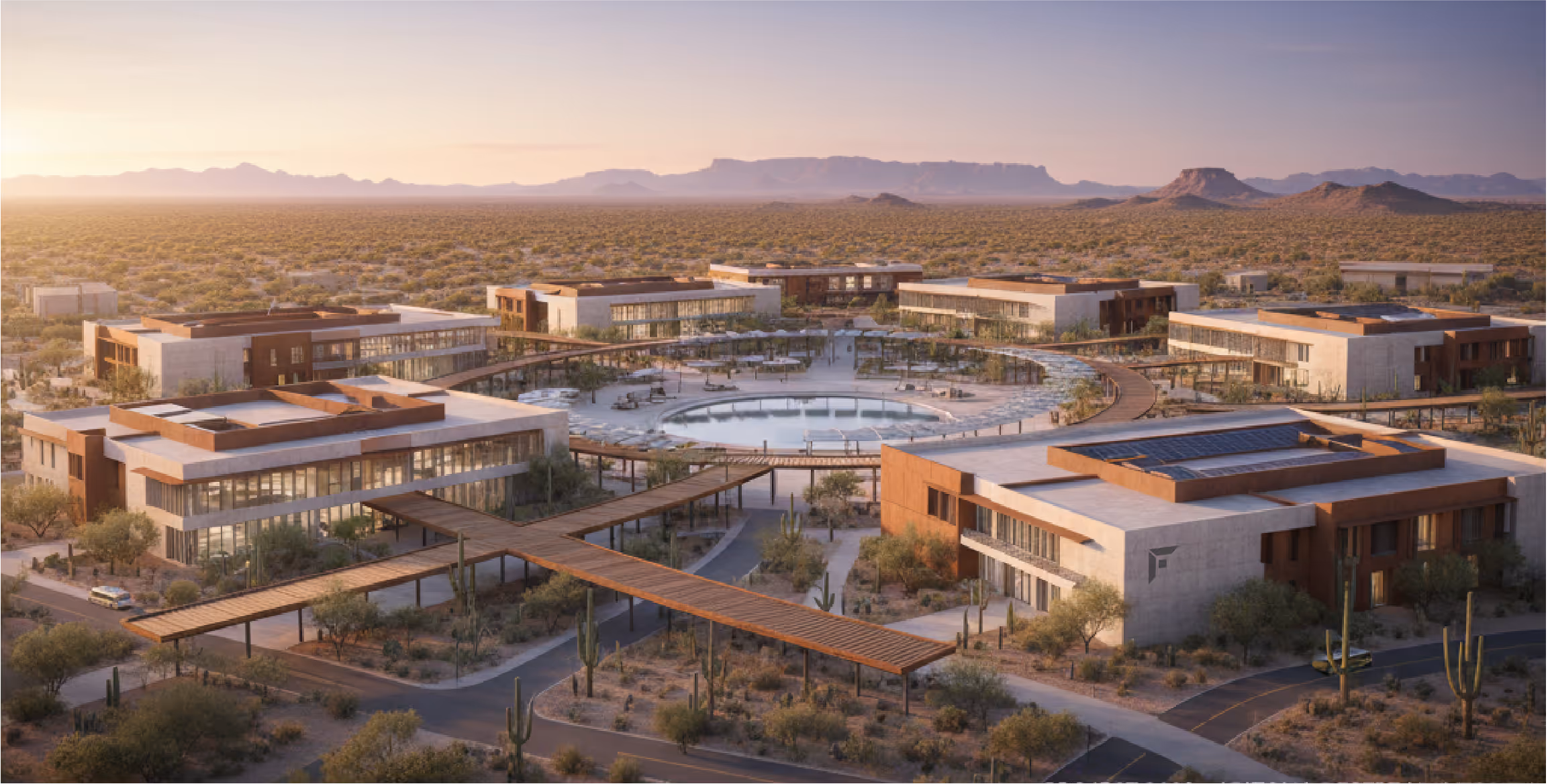Aerial view of a modern desert campus with six rectangular buildings surrounding a circular central pool, connected by covered walkways amid cacti and desert vegetation at sunset.