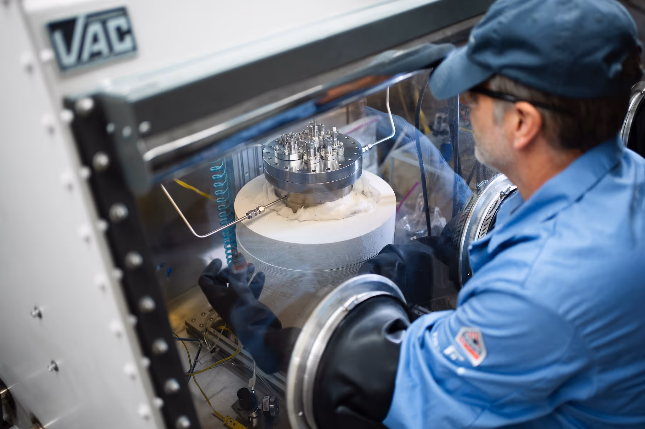 Scientist wearing black gloves and blue lab coat working with equipment inside a glass-enclosed glovebox in a lab.