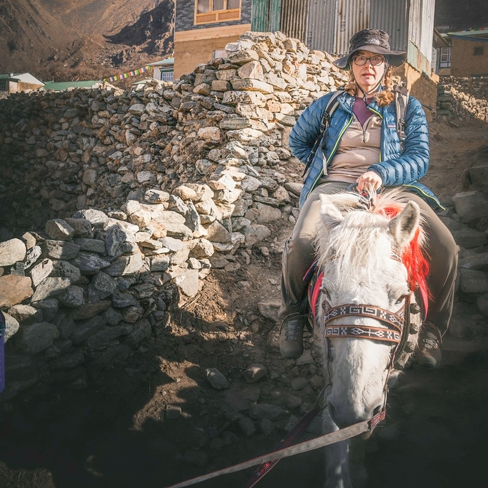 Person wearing a blue jacket and hat riding a white horse on a rocky mountain path beside stone walls and buildings.