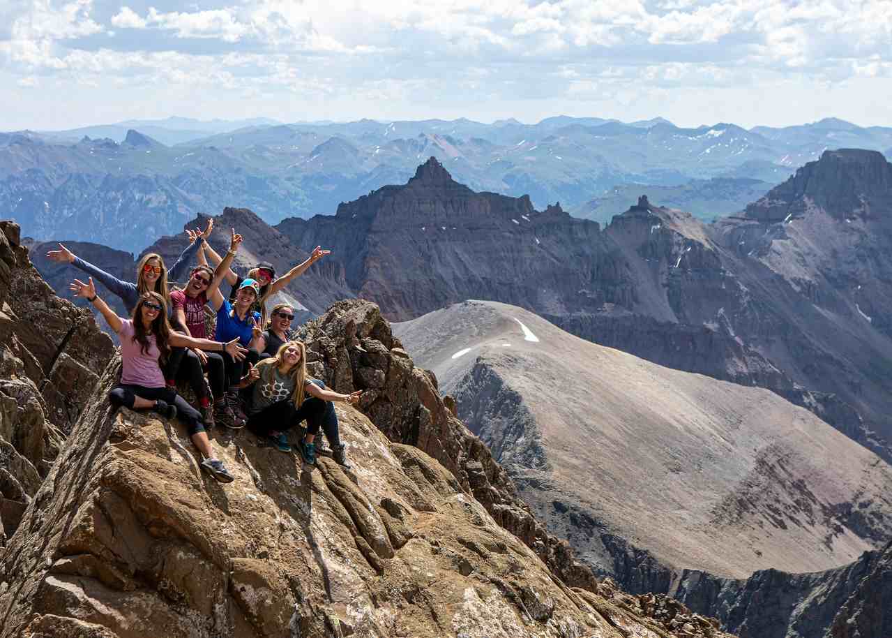 Group of seven hikers sitting on rocky mountain peak with expansive mountainous landscape in the background.