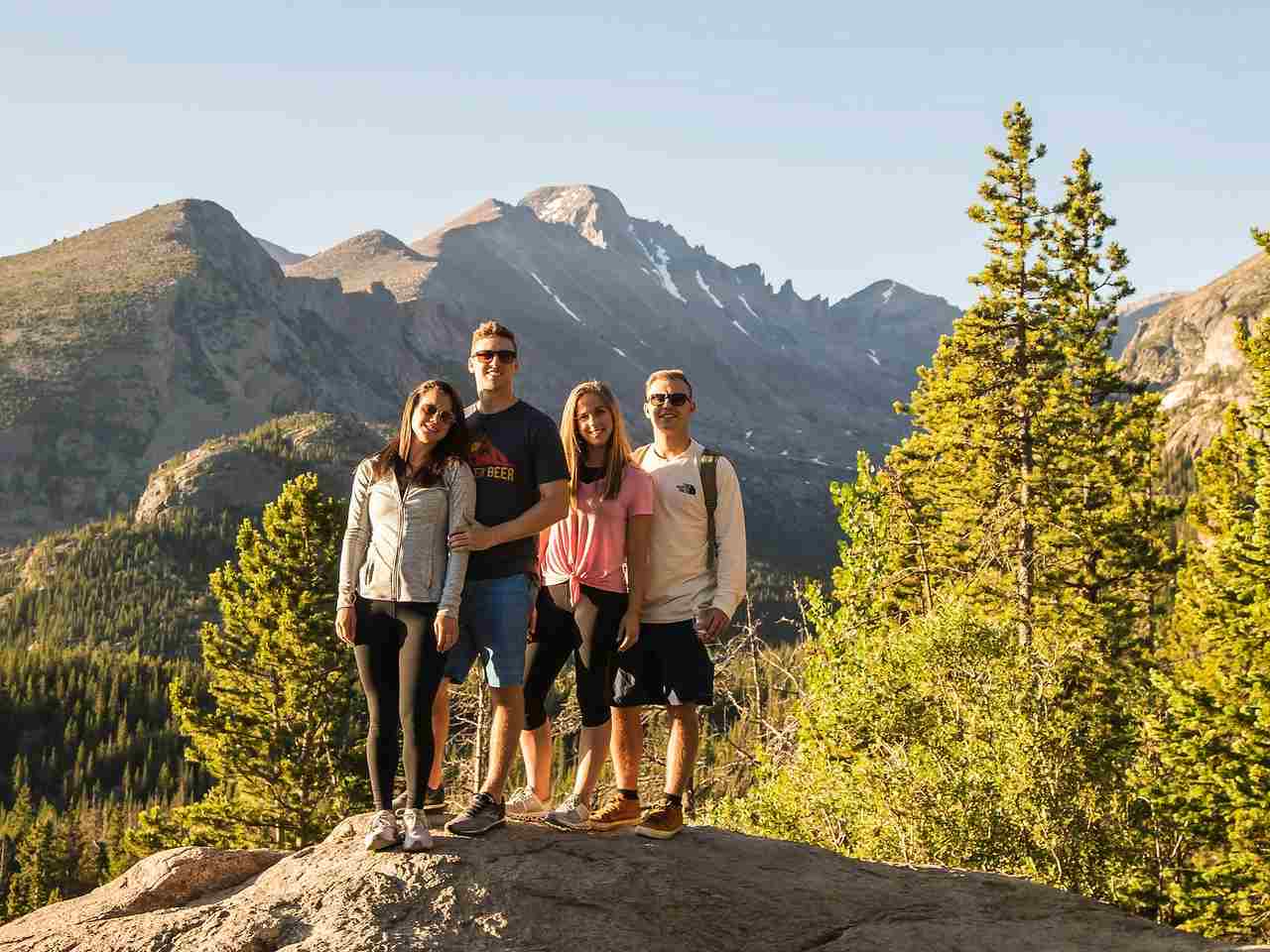 Four people standing on a rock with forest and mountain landscape in the background during daylight.