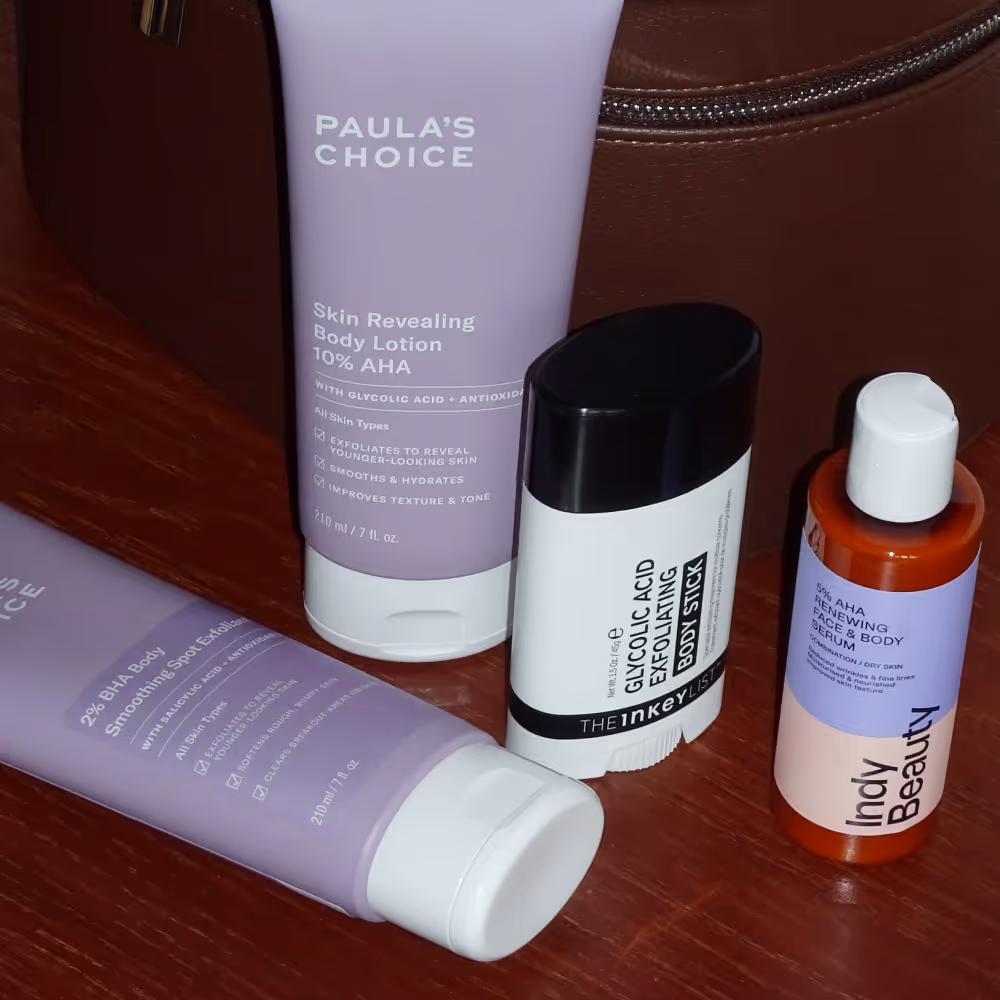 Tre produkter med AHA-syra som kan behandla keratosis pilaris: Paula's Choice Skin Revealing Body Lotion 10% AHA, The Inkey List Glycolic Acid Exfoliating Body Stick och den produkt som fick bäst i test i Glowaddictions stora test: Indy Beauty 5% AHA Renewing Face & Body Serum. Produkterna ligger och står på ett bord med en necessär i bakgrunden.