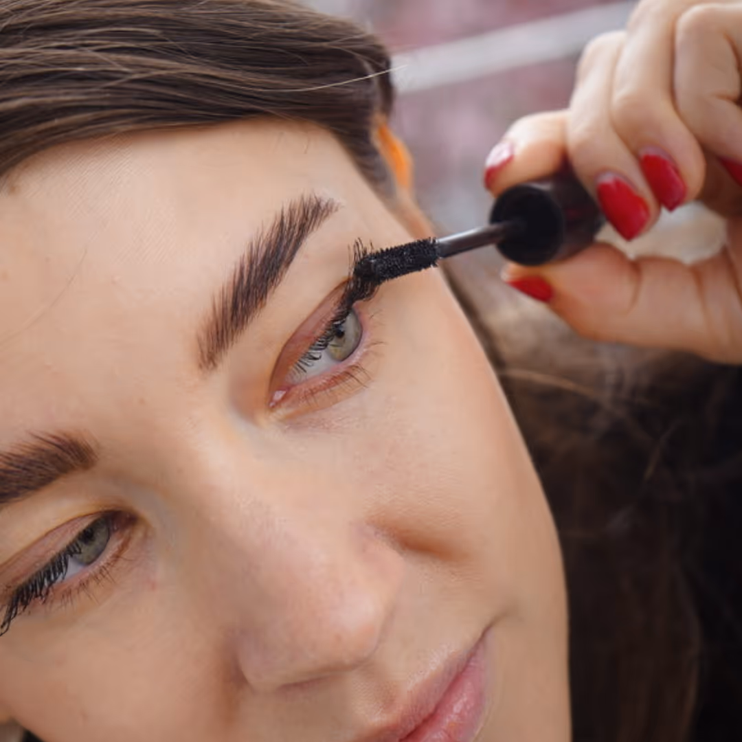 A woman with brown hair and red nail polish is applying mascara.