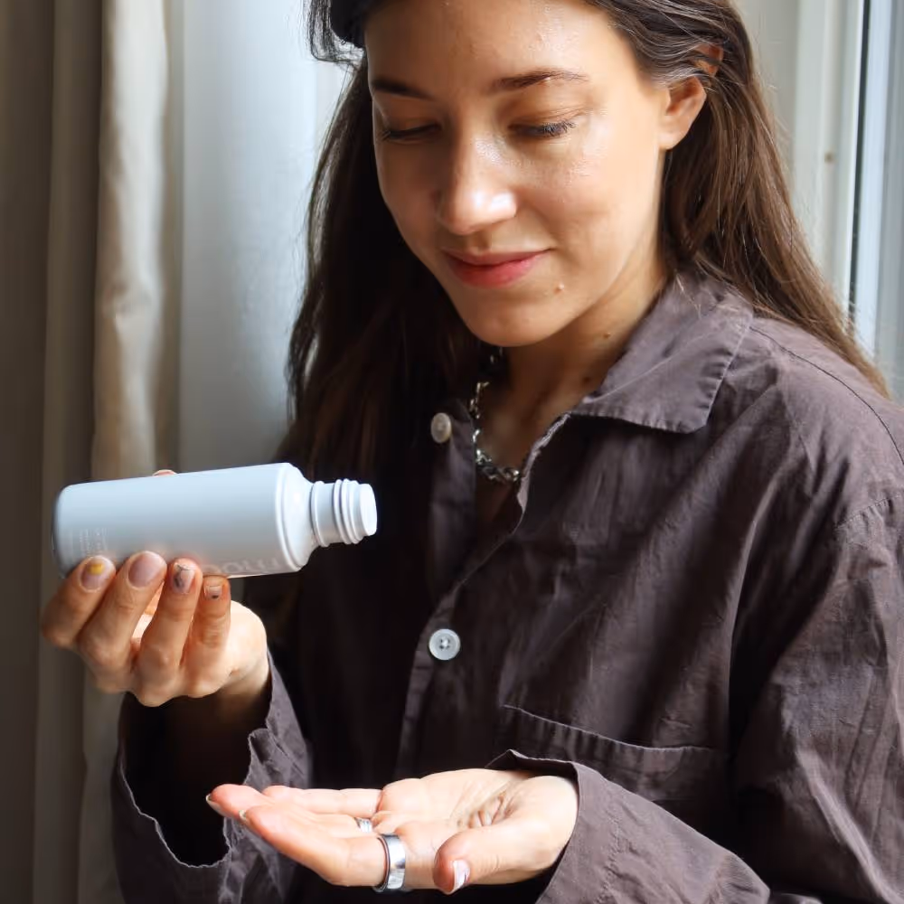Lina Livian håller i en förpackning med essencen Rhode Glazing Milk och ska hälla ut lite i handen, Lina har brunt hår och en brun skjorta, produkten syns tydligt i bild