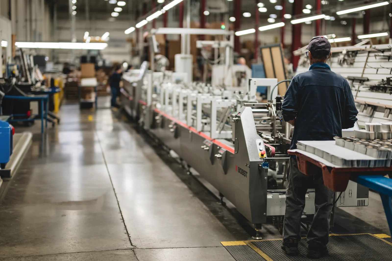 Operator at the delivery end of a Bobst folder-gluer line, with stacks of folded cartons on the conveyor in a folding carton plant