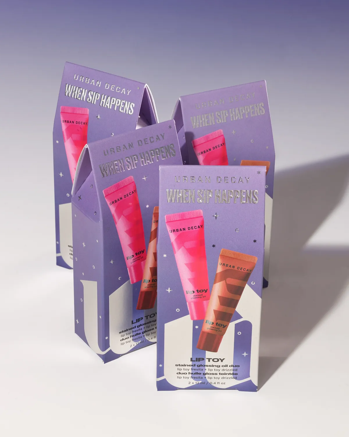 Four Urban Decay When Sip Happens Lip Toy duo cartons in lavender, with the wordmark and small stars in silver cold foil above printed images of pink and copper lip gloss tubes.