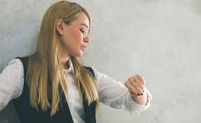 Young woman with long blonde hair wearing glasses and a black vest looking at her wrist.