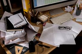Cluttered desk with stacks of papers, notebooks, eyeglasses, a keyboard, and stationery items.