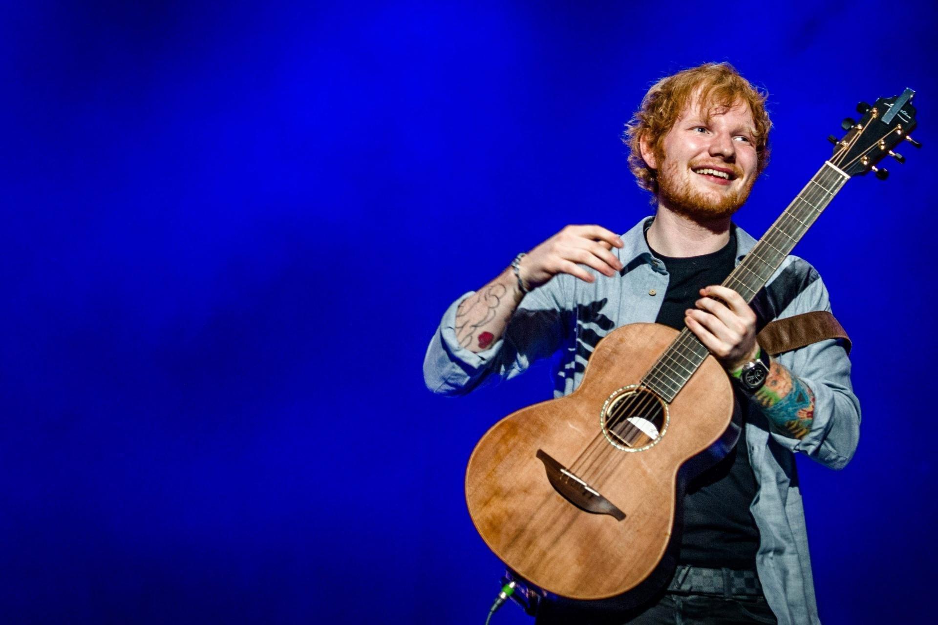 Ed Sheeran with tattoos playing acoustic guitar against a blue background on stage.