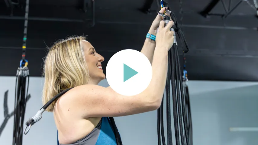 A woman smiles as she adjusts bungee harnesses in a studio with a modern, industrial vibe. The mood is energetic and focused.