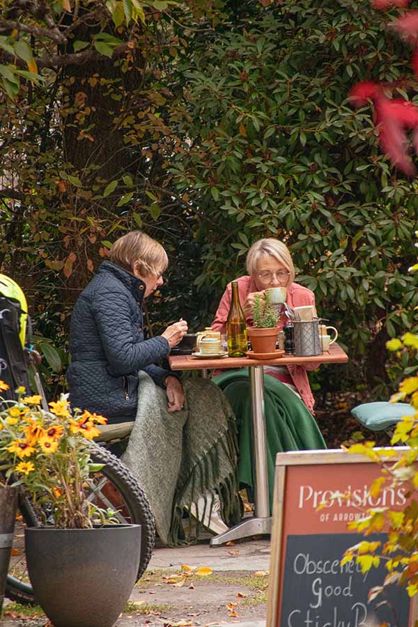 Dwie starsze osoby siedzące przy stole na świeżym powietrzu, udekorowanym kwiatami, rozmawiają ze sobą.