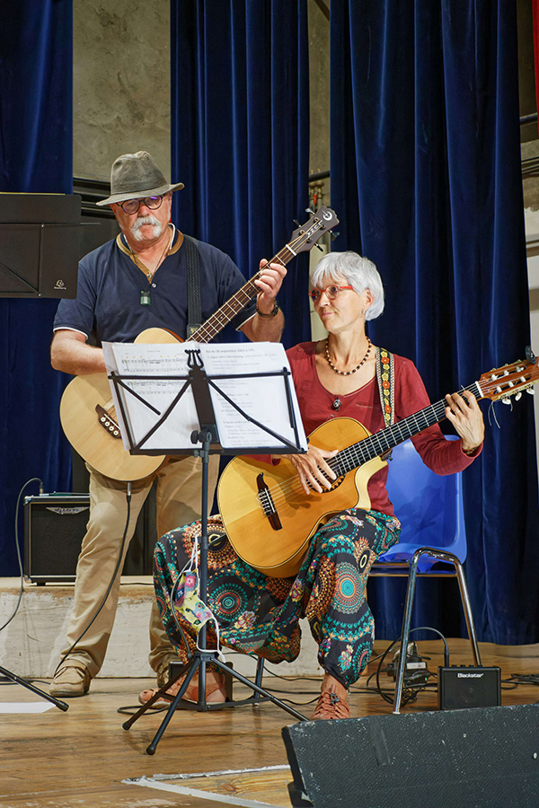 Dwóch muzyków na scenie – jedna osoba gra na gitarze, druga na skrzypcach, z mikrofonami i stojakami na nuty.