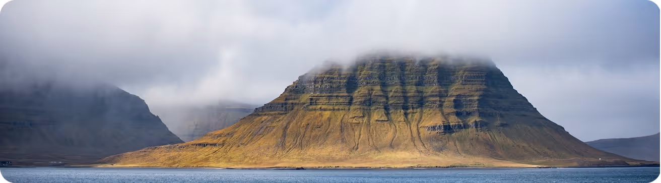 Góra o płaskim szczycie, częściowo zasłonięta nisko wiszącymi chmurami, otoczona wodą i odległą doliną.
