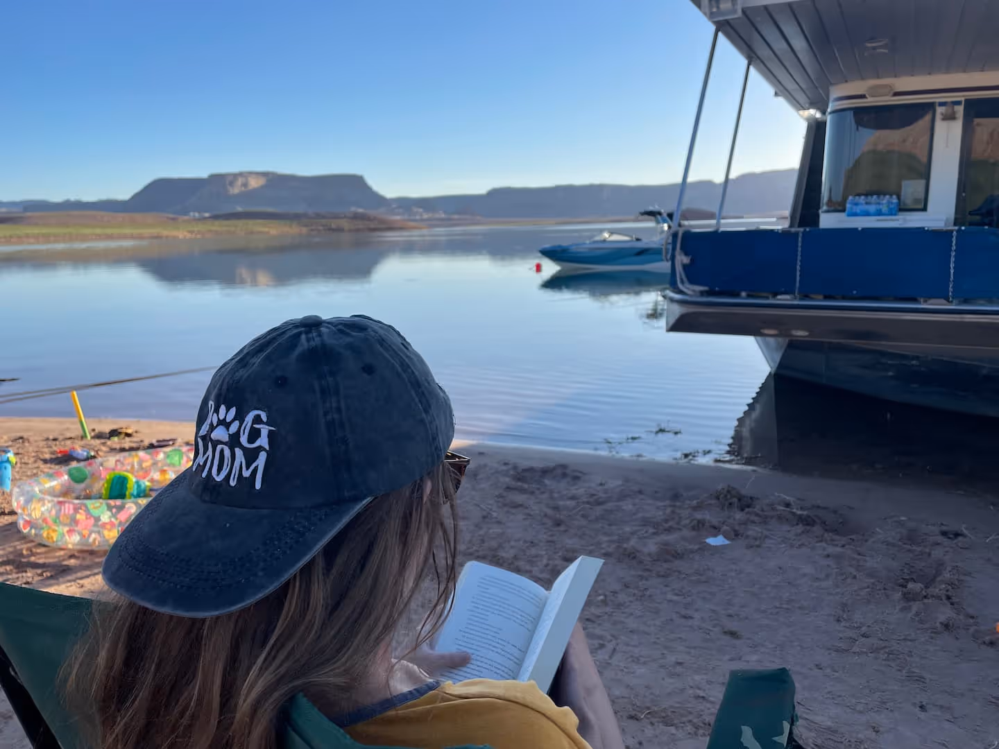 Woman wearing a “Dog Mom” hat sitting by the shore, reading a book, with boats docked on a calm lake in the background.