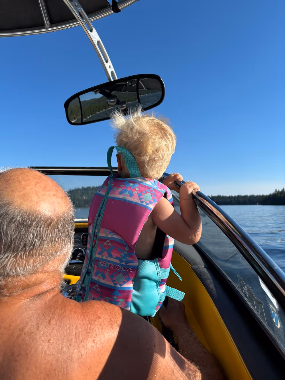 Man and child wearing life jackets on a boat, with the child sitting on the bow rail and looking out at the water on a sunny day.