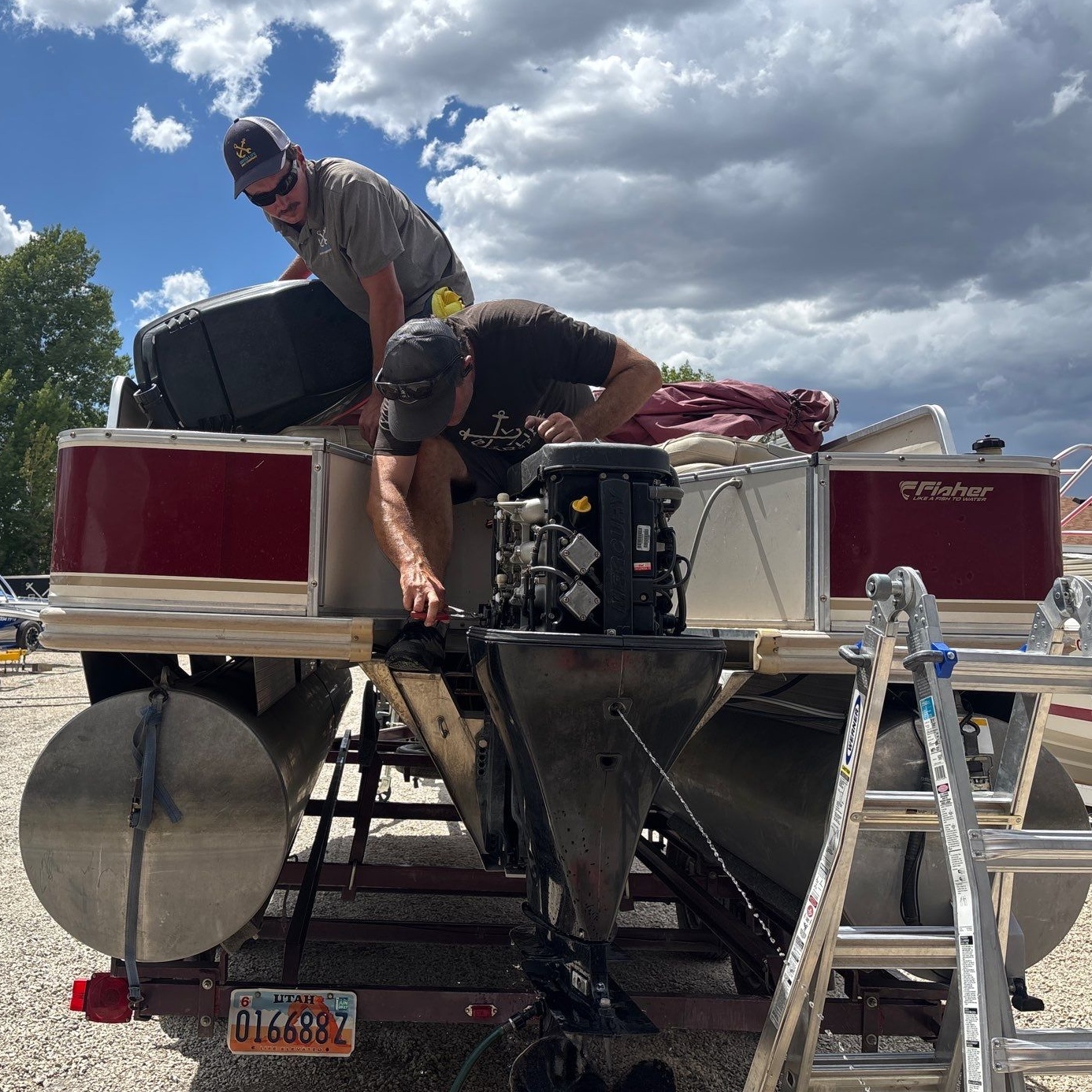 Two men working on an outboard engine on a pontoon boat on a trailer in a suburban neighborhood, discussing the boat’s condition in the warm evening sunlight with patches of melting snow on the lawn.
