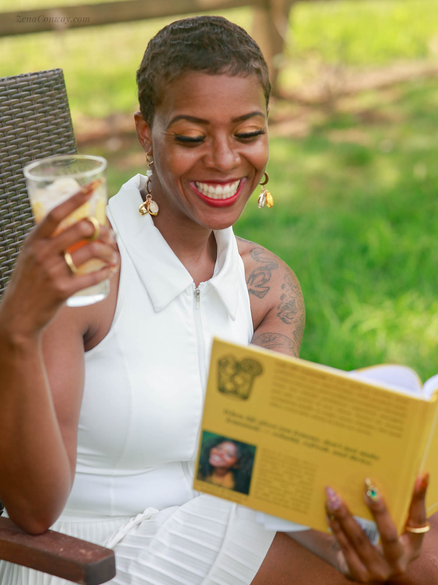 Zena C. smiling brightly while sitting outdoors enjoying her book "Laid Off Lemonade" and holding a refreshing glass of lemonade. 