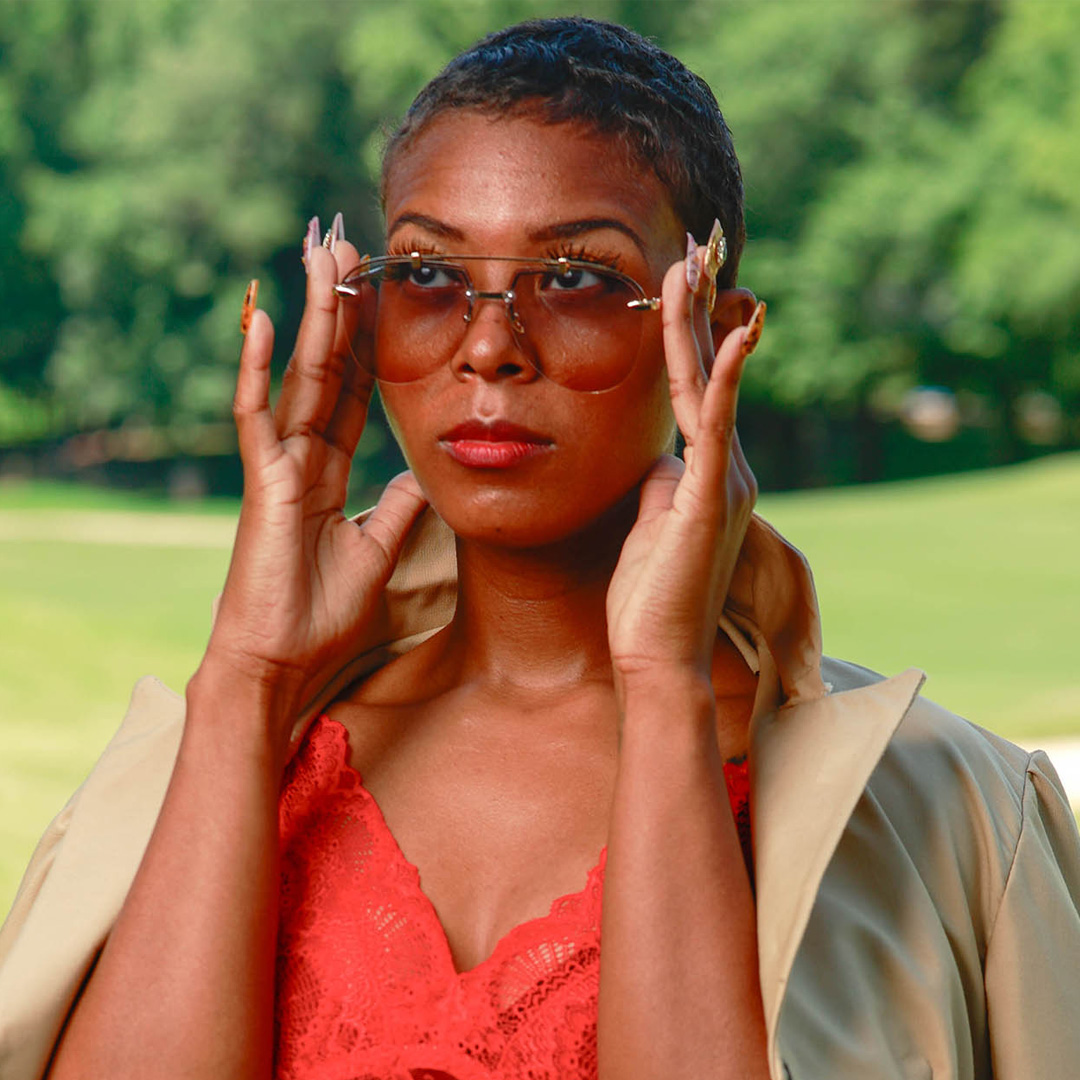 Zena C. in a bright red lace top with a tan blazer adjusting her translucent shades while looking serenely into the distance. 