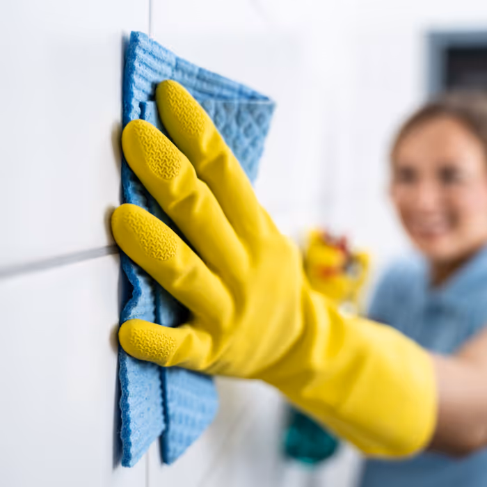 Person wearing yellow gloves cleaning a white tile wall with a blue cloth, representing reliable and detailed house cleaning services.