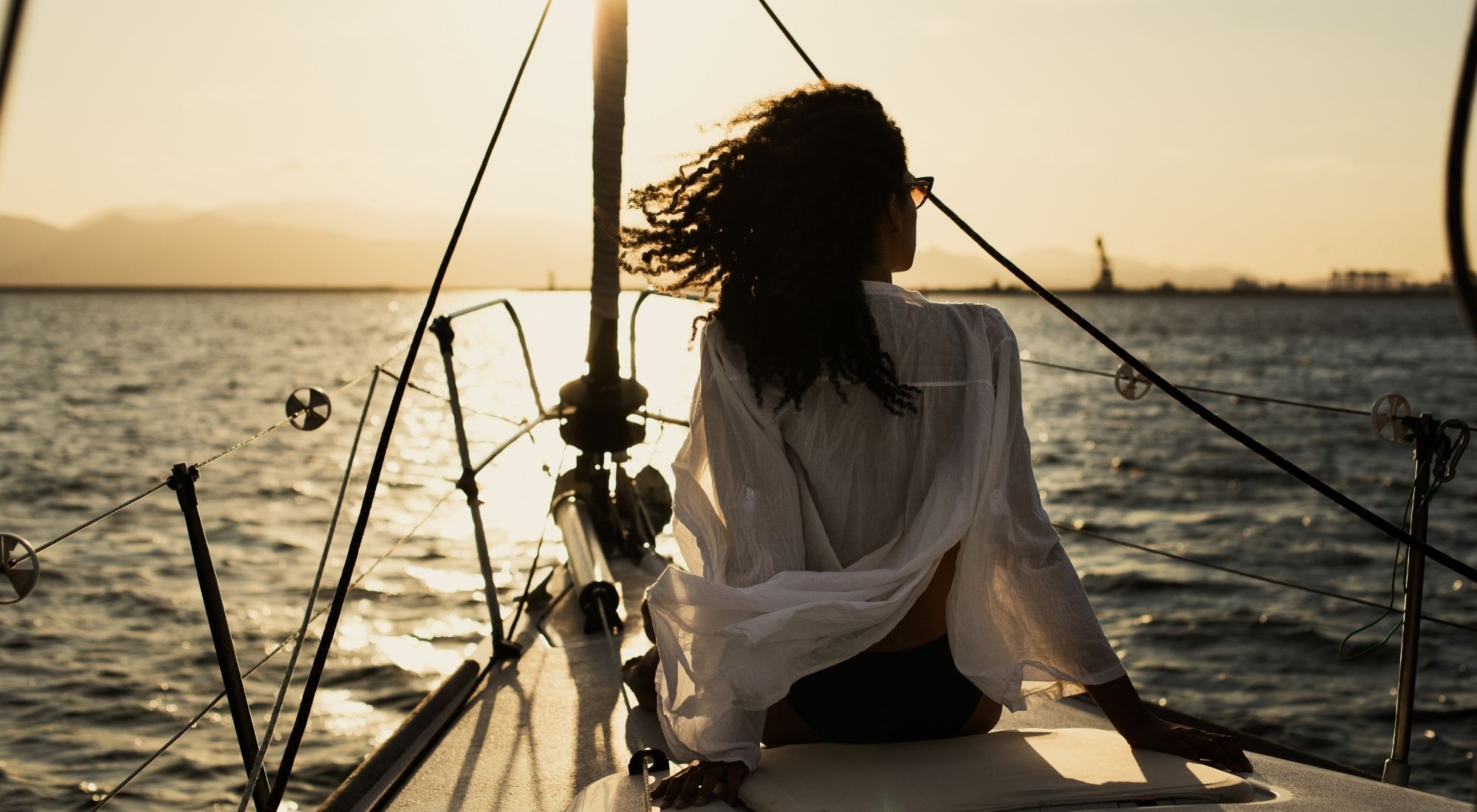 From behind, a woman sitting on bow of sailboat in a bay enjoying the sunset.