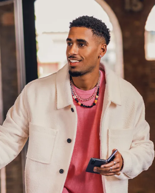 Young man wearing a white jacket and pink shirt holding a phone, smiling and looking to the side indoors.