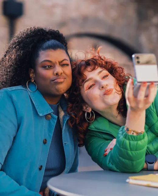 Two women sitting at a table taking a selfie; one making a funny face, the other puckering lips.
