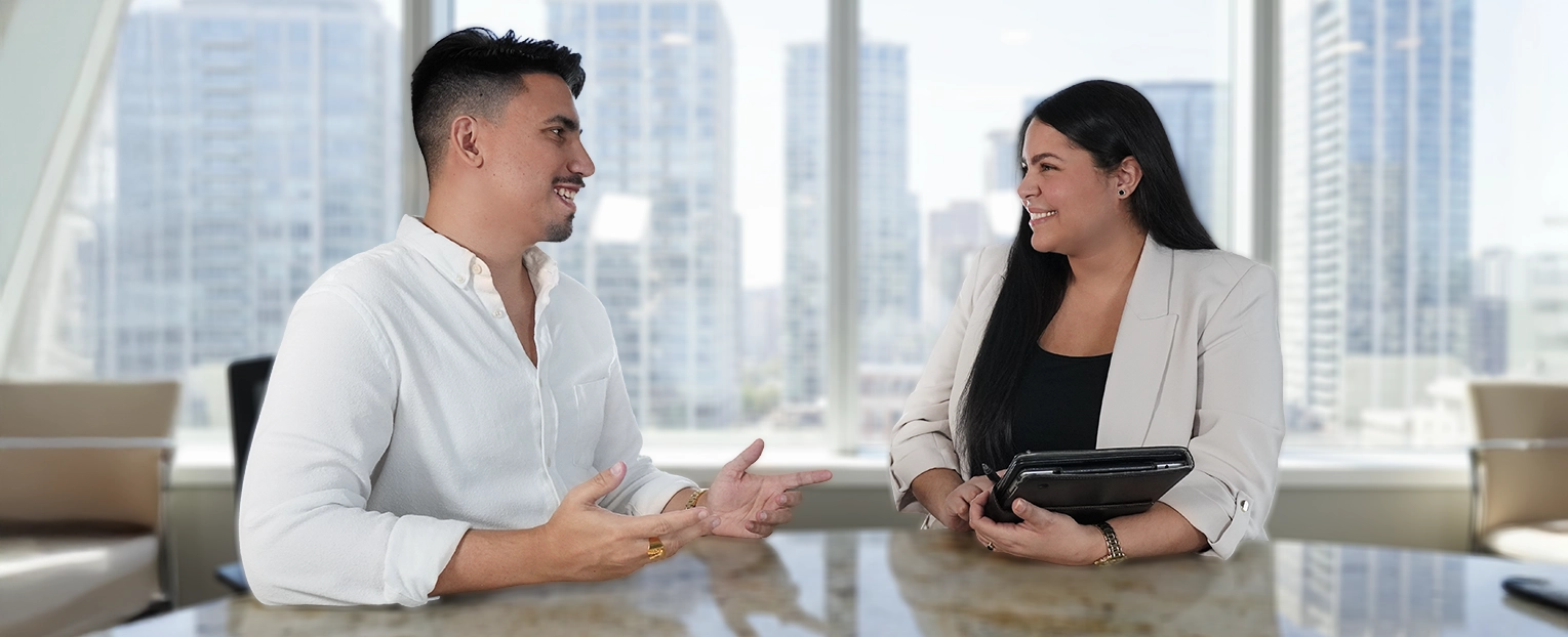 Dioni and Milfi, co-founders of Over the Fold, smiling and conversing across a table in a modern office with city buildings visible through large windows.