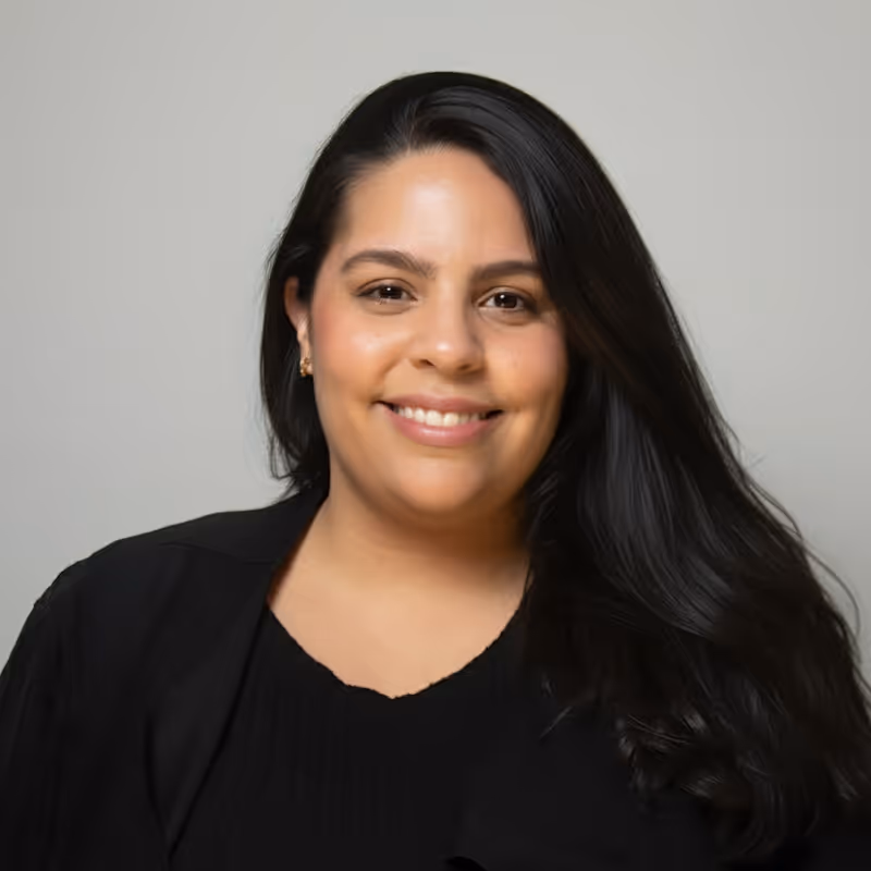 Smiling woman with long black hair wearing a black top and black jacket against a plain background.