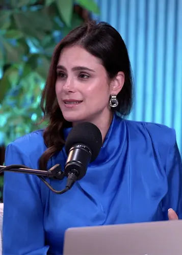 Woman in a blue blouse speaking into a microphone with a blurred green leafy background.