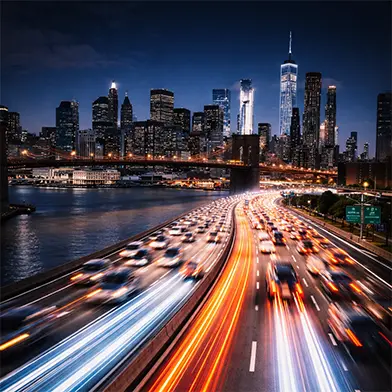 Nighttime view of busy highway with light trails leading to the illuminated Manhattan skyline and Brooklyn Bridge.