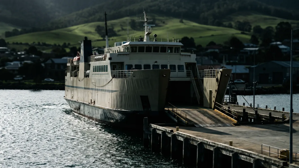 A passenger and freight ferry berthed at a river wharf with green hills behind.
