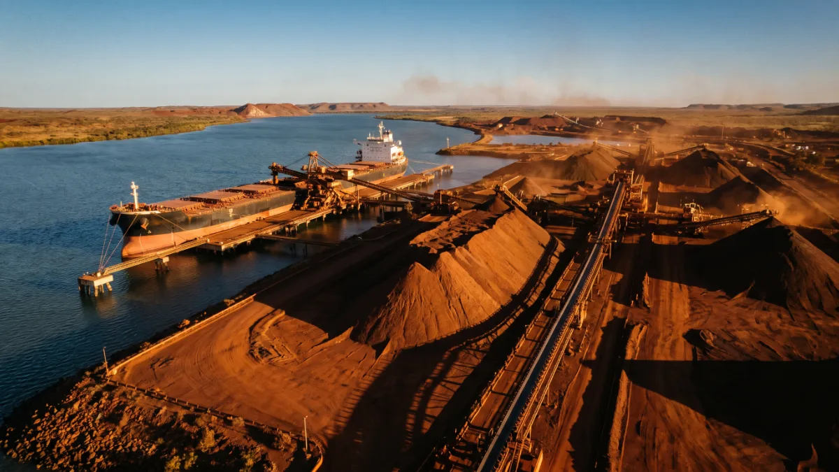 A bulk carrier ship berthed beside red iron ore stockpiles and loading conveyors at a remote Pilbara port at golden hour.