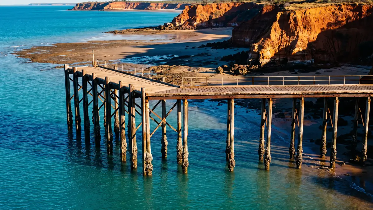 A timber jetty on pylons over turquoise water, with red coastal cliffs and a tidal sand and mudflat beach.