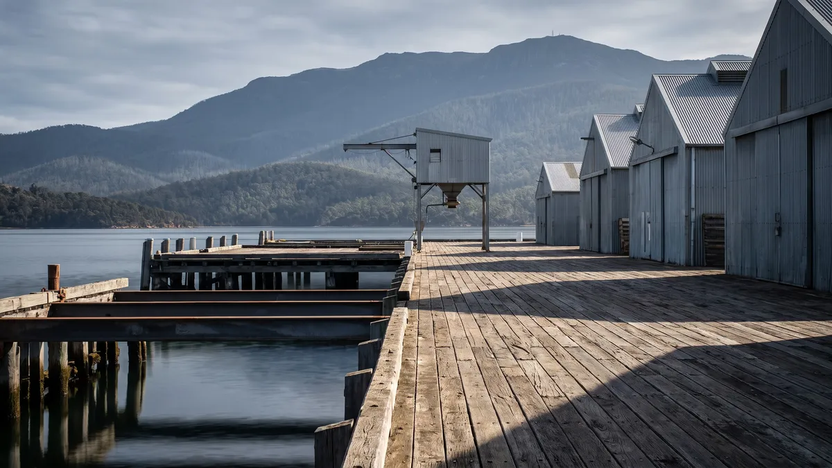 A timber wharf with corrugated cargo sheds and a small loading gantry, calm harbour water and forested mountains behind.