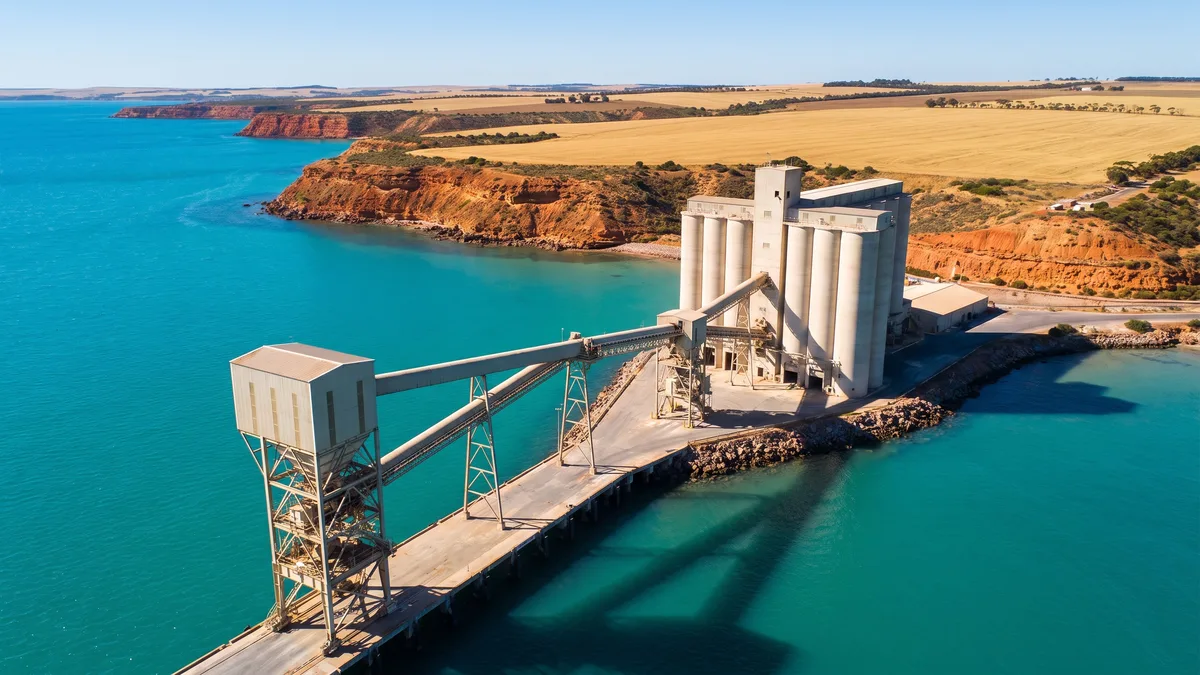 Grain silos and a conveyor loader on a wharf, with copper-coloured coastal cliffs and golden farmland behind turquoise water.